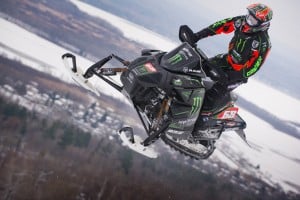A snowmobile racer in a black and green outfit is airborne while riding a snowmobile featuring multiple sponsor logos. The background showcases a snowy landscape with trees and a distant view of a wintery village.