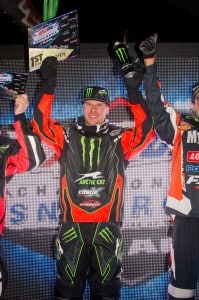 A snowmobile racer in a red and black racing suit stands on a podium holding a plaque above his head. He is flanked by two other racers on either side, also holding plaques. The background features a large, illuminated screen with logos and text.