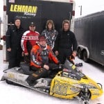 A group of five people poses in front of a Lehman Racing trailer. One person is seated on a yellow snowmobile with racing decals, while the other four stand behind. Snow covers the ground, and various trailers are visible in the background.