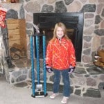 A young girl stands in front of a stone fireplace, smiling and wearing a red-orange jacket with a colorful pattern. She has gloves on and is next to a tall trophy with blue accents. There is a Christmas stocking hanging on the left side of the fireplace.