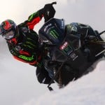 A snowmobile rider wearing a colorful helmet and black racing gear with green and orange accents navigates through a snowy terrain. The snowmobile is decorated with various logos, including Monster Energy. Snow sprays around the vehicle as it speeds forward.