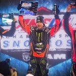 A snowmobile racer in a red and black Arctic Cat racing suit celebrates on a podium holding up a 1st place certificate and a drink can. The racer is flanked by two other racers in similar suits holding their certificates.