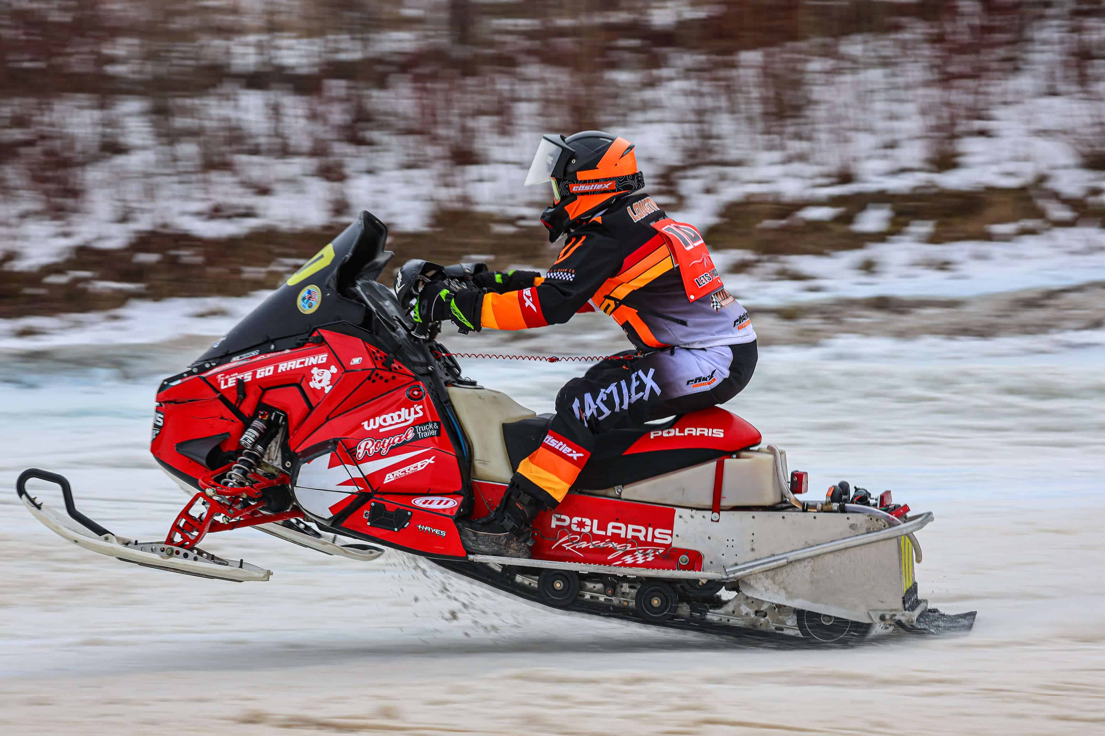 A person in a colorful racing suit and helmet rides a red snowmobile fast across a snowy landscape. The snowmobile has various logos and branding. The background features patches of snow and bare trees.