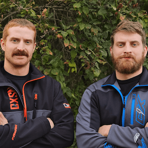 Two men with beards and mustaches stand with arms crossed, wearing jackets with logos. They are outdoors, in front of leafy greenery, looking at the camera.
