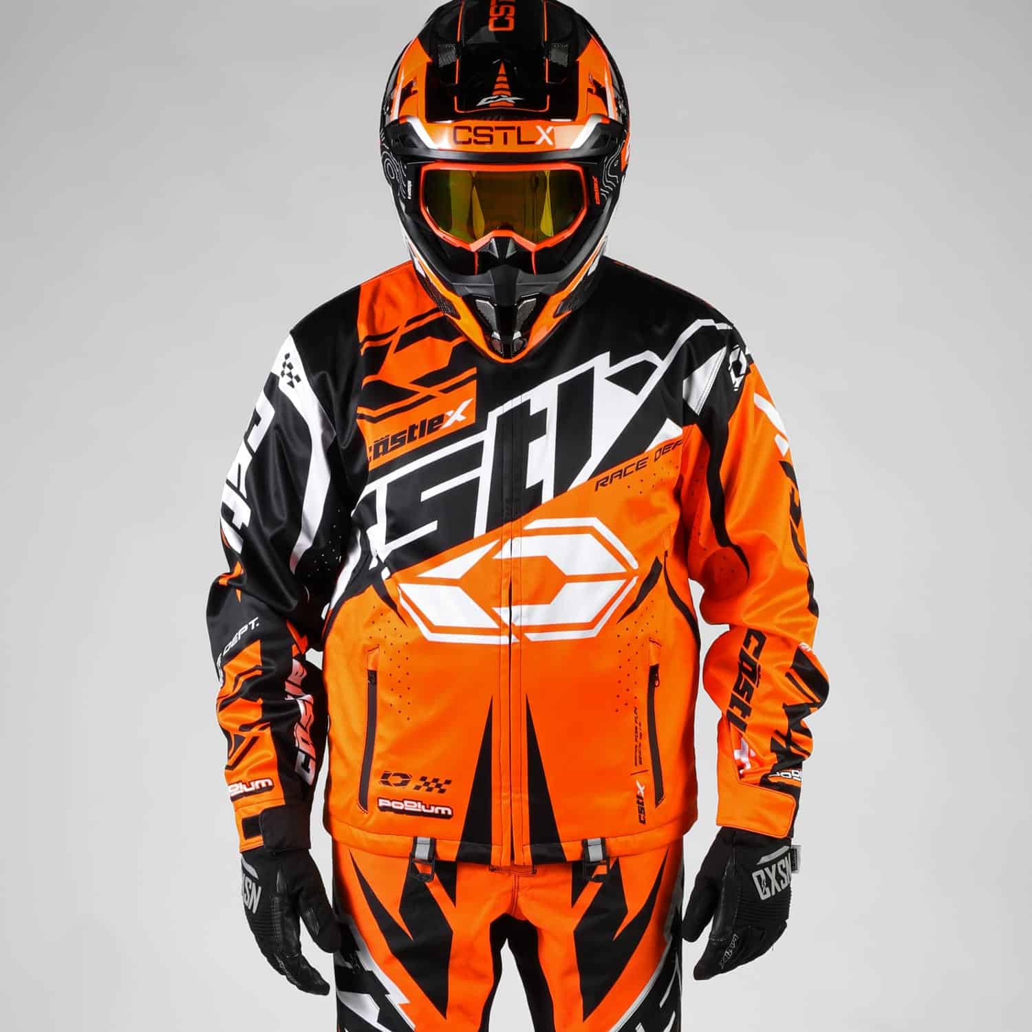 A person wearing the Men’s Podium Jacket—a bright orange, black, and white racing jacket with bold graphics—along with a matching helmet, gloves, and goggles, stands against a plain light gray background.